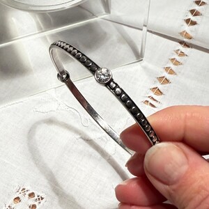 May include: A silver bangle bracelet featuring a central clear gemstone and small, dark embellishments. The bracelet is displayed against a white backdrop, highlighting its intricate design. This is a piece of jewellery.