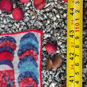 May include: A red, blue, and white patterned fabric square with a red border. The fabric is lying on a bed of small gray stones. A yellow measuring tape is visible in the background.