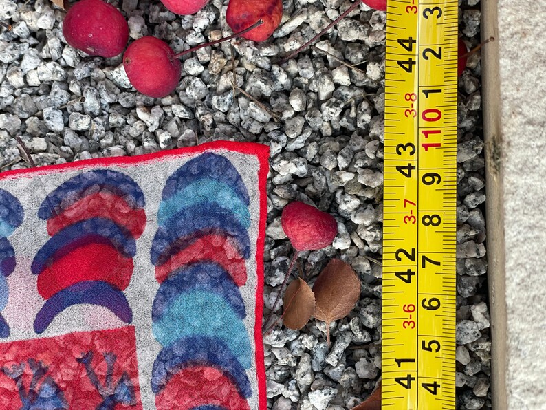 May include: A red, blue, and white patterned fabric square with a red border. The fabric is lying on a bed of small gray stones. A yellow measuring tape is visible in the background.