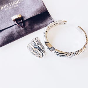 May include: A silver cuff bracelet and ring set with intricate etched designs and small gemstones. A dark gray pouch with the text "JOHN HARDY EST. BALI 1975" is also present. The jewelry is displayed on a white surface.