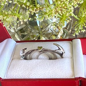May include: A silver-toned ring featuring a wrap-around design that resembles two hands meeting at the center, accented with a small, singular green heart-shaped detail. The ring is displayed inside a red velvet-lined jewelry box.