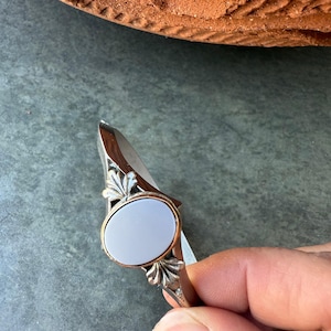 May include: A silver-toned ring with an oval, pale blue stone set in a gold-toned bezel. The ring features ornate leaf-like designs on the sides. The ring is held by a hand against a gray surface.