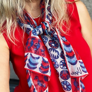 May include: A white silk scarf with a red, blue, and white geometric pattern. The scarf features stylized birds and circles. The scarf is tied around the neck of a person wearing a red sleeveless top.