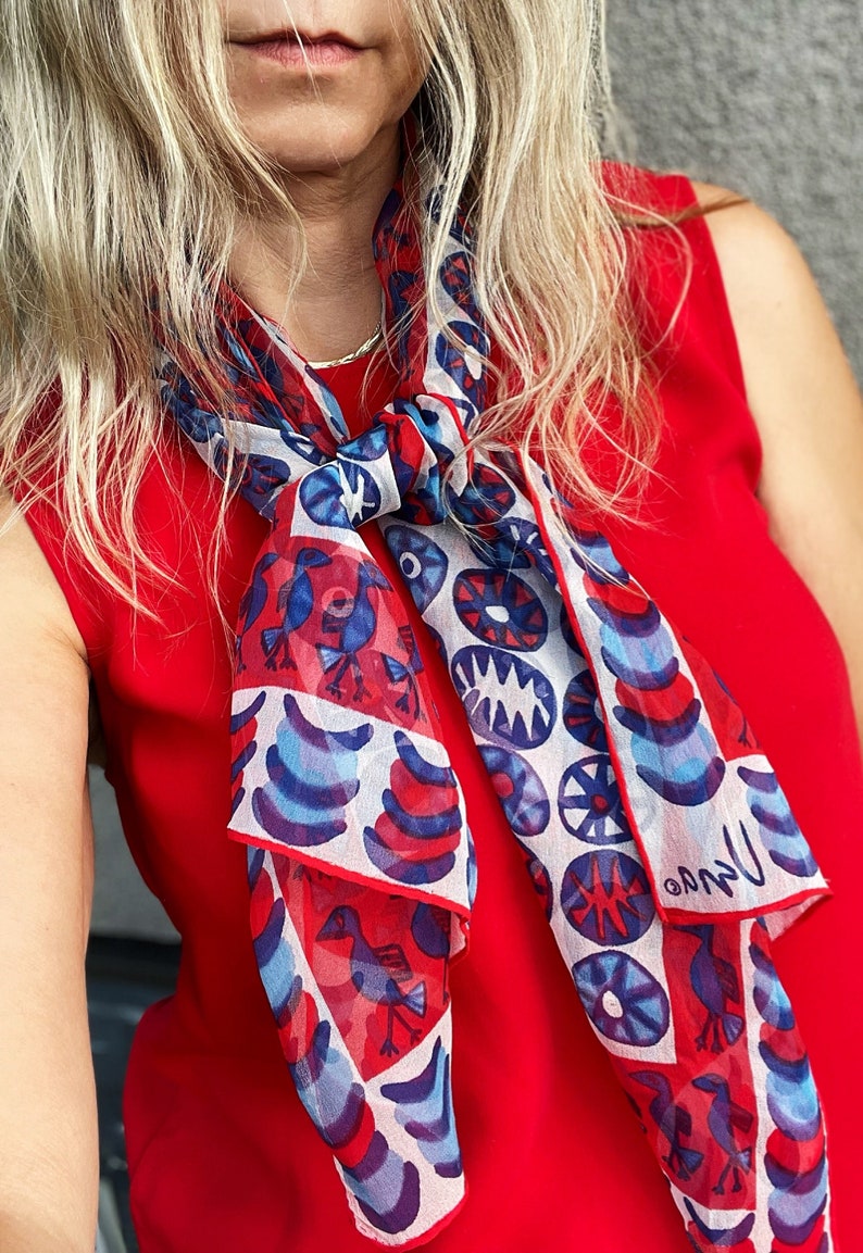 May include: A white silk scarf with a red, blue, and white geometric pattern. The scarf features stylized birds and circles. The scarf is tied around the neck of a person wearing a red sleeveless top.