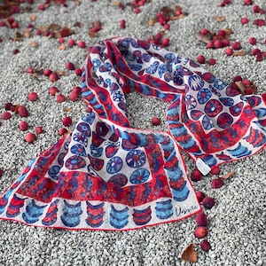 May include: A white silk scarf with a red and blue geometric pattern. The scarf is folded in half and lying on a bed of small red berries.