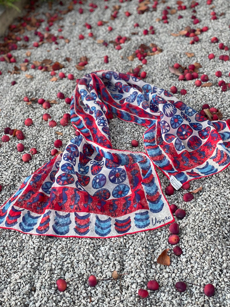 May include: A white silk scarf with a red and blue geometric pattern. The scarf is folded in half and lying on a bed of small red berries.