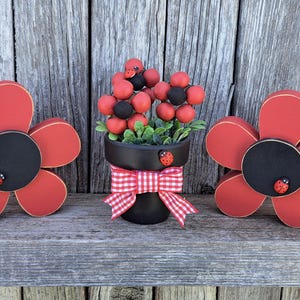 May include: Wooden flower decorations with red petals, black centers, and ladybug accents. A black flower pot with a red and white checkered bow holds a floral arrangement. The items are displayed on a wooden surface.