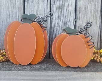 May include: Two orange wooden pumpkin decorations with green leaf accents and wire tendrils. The pumpkins are displayed on a rustic wooden surface, evoking a fall or harvest theme. Decorative fall accents are in the background.