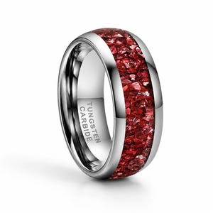 May include: A silver tungsten carbide ring with a vibrant red inlay. The ring has a polished, reflective surface and features the text "TUNGSTEN CARBIDE" on the inside. The red inlay is made of small, irregular pieces.