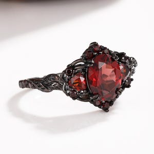 May include: A black metal ring featuring a large oval-cut deep red gemstone at the center, flanked by smaller triangular red stones. The band is detailed with a raised leaf pattern, and the setting is accented with tiny black round stones.