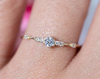 May include: A delicate gold ring featuring a central round diamond and smaller accent stones. The ring's band has a slender design with a floral-inspired pattern. The ring is shown on a person's finger, with a blurred pink background.