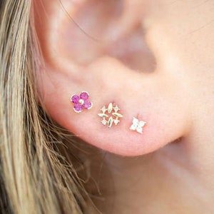 May include: A close-up of a person's ear with three small earrings. The earrings are a pink flower, a gold geometric design, and a small diamond.