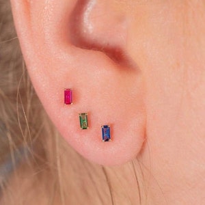 May include: Three small gold earrings with rectangular gemstones. The earrings are in a row, with a red gemstone on the left, a green gemstone in the middle, and a blue gemstone on the right.