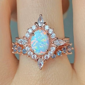 May include: A rose gold ring with a large oval opal surrounded by smaller clear stones. The ring has a delicate design with a pointed top and a band of smaller blue stones.