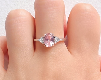 May include: A silver ring featuring a large, oval-cut pink gemstone as the centerpiece, flanked by smaller, round, clear gemstones on each side. The ring is set on a delicate band, showcasing a classic and elegant design.