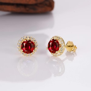 May include: A pair of gold earrings with a red gemstone surrounded by a halo of smaller white gemstones.