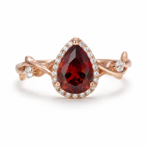 May include: A rose gold ring featuring a pear-shaped deep red center stone surrounded by a halo of small white stones. The band is designed with a textured, branch-like motif that includes small leaves and additional white stones on each side.