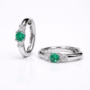 May include: Two silver hoop earrings, each featuring a vibrant green emerald center stone flanked by two smaller, clear gemstones. The earrings are set against a white background, highlighting their elegant design.