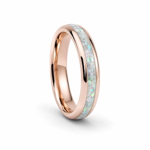 May include: A rose gold band ring featuring a central inlay of crushed white opal with iridescent flecks. The metal surface is polished and reflective, creating a smooth, rounded profile that frames the textured, shimmering stone inlay.
