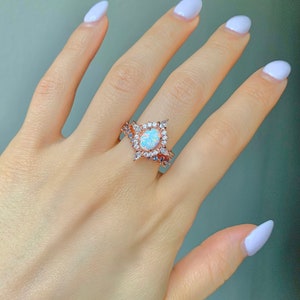 May include: A rose gold ring with a large oval opal stone surrounded by smaller clear stones. The ring is on a hand with white nail polish.