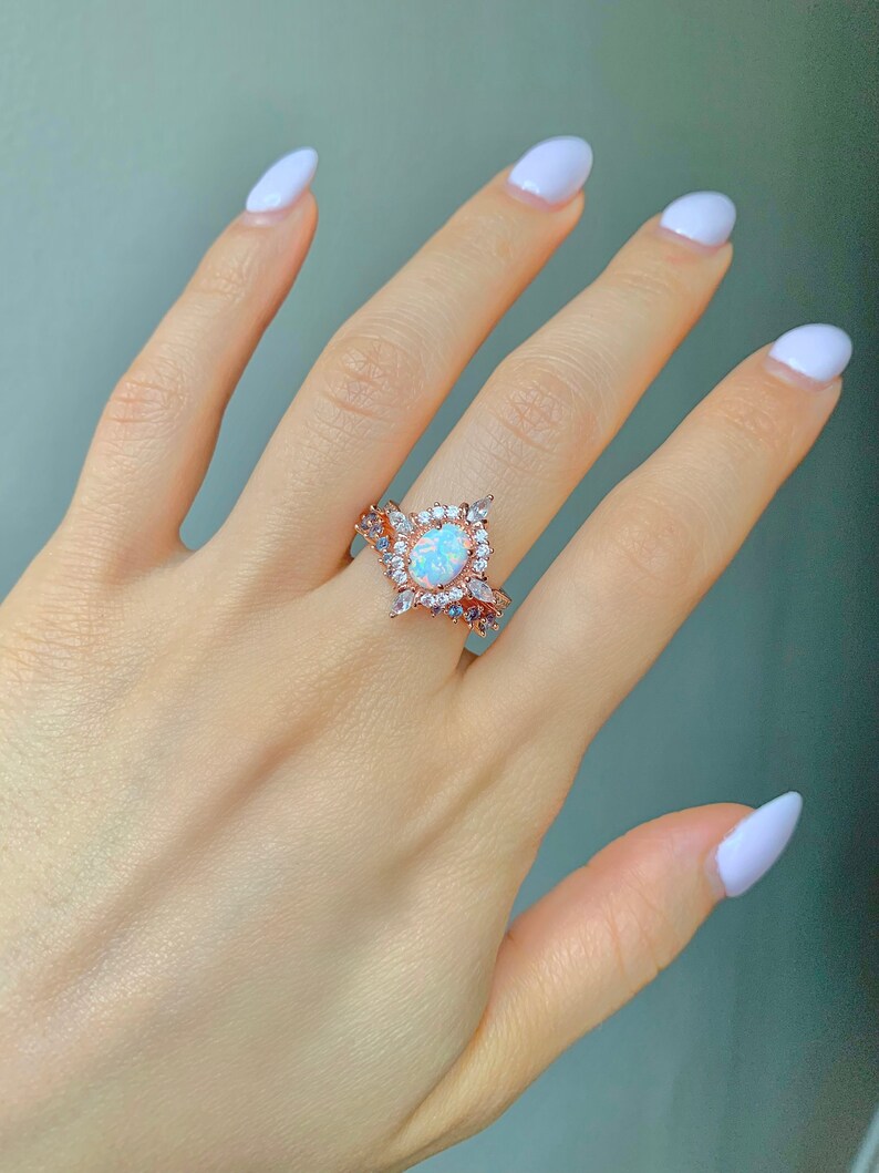 May include: A rose gold ring with a large oval opal stone surrounded by smaller clear stones. The ring is on a hand with white nail polish.