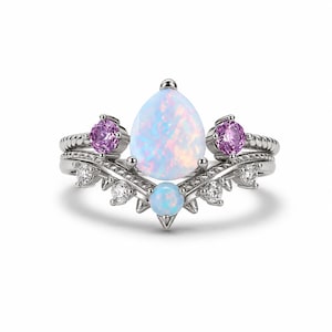 May include: A silver-toned ring featuring a central pear-shaped iridescent opal, two round pink stones, a smaller round opal at the base, and four small clear stones set along a chevron-shaped band with a textured, rope-like metal finish.