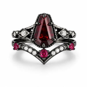 May include: A black metal ring set featuring a central coffin-shaped deep red gemstone. The band is accented with small clear white stones and round red stones arranged in a chevron pattern, creating a layered look with dark metal prongs.
