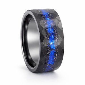 Puede incluir: Un anillo negro con una superficie facetada y una incrustación azul y plateada. El anillo tiene una banda ancha y un interior pulido. La incrustación azul tiene una apariencia texturizada, creando un contraste llamativo.