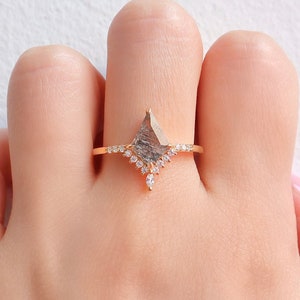 May include: A gold ring with a large, gray, faceted gemstone in a kite shape. The gemstone is surrounded by a halo of smaller, clear gemstones. The ring is on a finger.