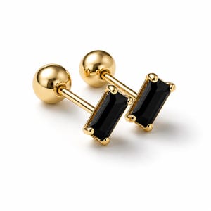 May include: A pair of gold stud earrings featuring a rectangular black baguette-cut gemstone held by four prongs. Each earring has a smooth, polished gold ball backing attached to a straight post, creating a clean and reflective metallic finish.