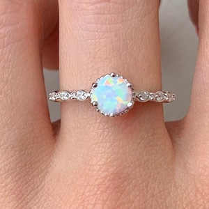 May include: A silver ring with a white opal gemstone surrounded by smaller clear gemstones. The ring is on a finger.