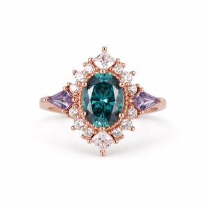 May include: A rose gold ring featuring an oval teal gemstone at the center, surrounded by a halo of small white and clear stones. Two triangular purple stones flank the center stone. The ring has a vintage, art deco style.