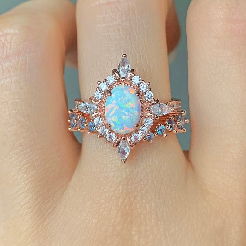 May include: A rose gold ring with a large oval opal surrounded by smaller clear stones. The ring has a delicate design with a pointed top and a band of smaller blue stones.