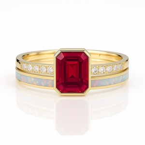 May include: A gold ring set featuring a large, rectangular, red gemstone in a bezel setting. The set includes a band with small, clear gemstones and another band with a white opal-like inlay. The rings are displayed on a white surface.
