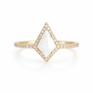 May include: A gold ring featuring a white, kite-shaped opal center stone, surrounded by a halo of small, clear gemstones. The band is also adorned with small gemstones. The ring is set against a white background.