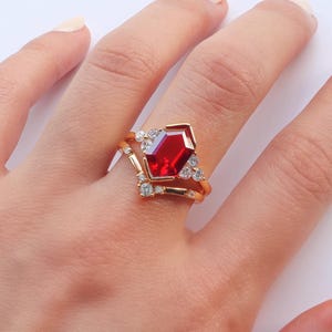 May include: A gold ring with a large, red, octagonal gemstone surrounded by smaller, clear gemstones. The ring is set on a finger.