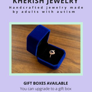 May include: A blue velvet ring box with a gold ring with a purple gemstone inside. The box is sitting on a wooden surface.  "KHERISH JEWELRY Handcrafted jewelry made by adults with autism GIFT BOXES AVAILABLE You can upgrade to a gift box or gift wrap for extra flare or a special occasion."