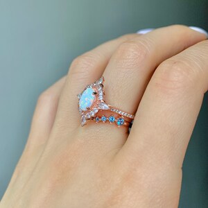 May include: A rose gold ring with a large oval opal surrounded by smaller clear stones. A smaller rose gold band with blue stones is worn on the finger below the larger ring.