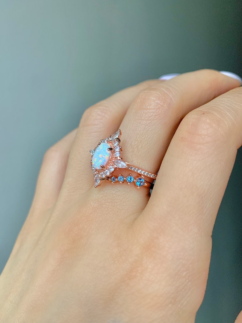 May include: A rose gold ring with a large oval opal surrounded by smaller clear stones. A smaller rose gold band with blue stones is worn on the finger below the larger ring.