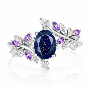 May include: A silver ring featuring a large, oval, dark blue gemstone with sparkling inclusions. The ring is adorned with leaf and amethyst accents. The leaves and amethyst stones are arranged around the central stone, creating a nature-inspired design.