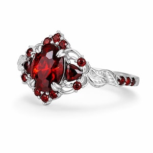 May include: A silver ring featuring a large oval-cut deep red garnet as the centerpiece, surrounded by smaller round and pear-cut red garnet stones. The band is crafted with a detailed leaf-like pattern and accented with additional small red stones.