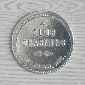 May include: A silver-colored metal token with the words "CLUB CHARMING" and "DELAVAN, WIS." embossed on it. The token has a decorative border and a cross and leaf design.