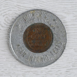 May include: A silver token with the text "BRUNER'S PARK FREE AND CAVE SAN ANTONIO, TEXAS" around the edge. In the center is a copper-colored coin with the text "ONE CENT UNITED STATES OF AMERICA".
