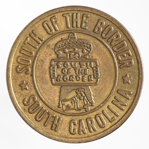 May include: A vintage, circular brass token with raised lettering and design. The text "SOUTH OF THE BORDER" arches around the top and "SOUTH CAROLINA" around the bottom. A central emblem reads "SOUTH OF THE BORDER" above a cartoon figure.