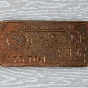 May include: A rectangular, bronze-colored metal bar with the text "YOUR KEY TO PROFITS" and "TOMORROW'S TECHNOLOGY TODAY." It features a key design and an illustration of machinery. The bar is marked with "275 TON" and a circular emblem.