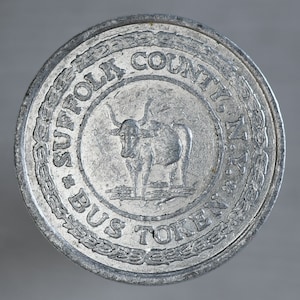 May include: A silver-colored Suffolk County, NY bus token. The token features an image of a bull in the center, surrounded by the words "SUFFOLK COUNTY, N.Y." and "BUS TOKEN". The token has a circular shape with a detailed border.