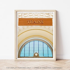 May include: A vintage photograph of a train station interior with a wooden sign that reads "TRAINS" hanging from the ceiling. The sign is above a large arched window with stained glass panels. The window is framed by a white archway and a yellow ceiling.