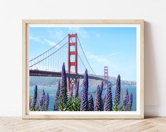 May include: A photograph of the Golden Gate Bridge in San Francisco, California. The bridge is red and has a blue sky and water in the background. There are purple flowers in the foreground.