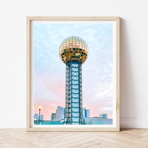 Puede incluir: Impresión enmarcada de la Sunsphere en Knoxville, Tennessee. La esfera dorada se asienta sobre una estructura alta de acero. El cielo es una mezcla de azul y rosa, con edificios de la ciudad al fondo. La impresión está en un marco de madera clara.