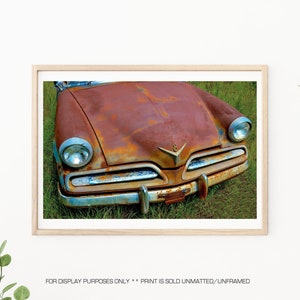 May include: Close-up of a rusty, vintage car with a chrome bumper and headlights. The car is parked in a field of green grass.